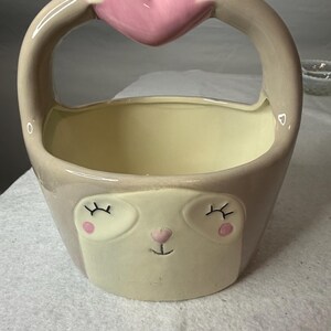 May include: A ceramic basket featuring a light beige sloth face with closed eyes, pink cheeks, and a small nose. The handle is formed by two arms reaching upward to hold a large, smooth, pink heart shape centered above the open container.