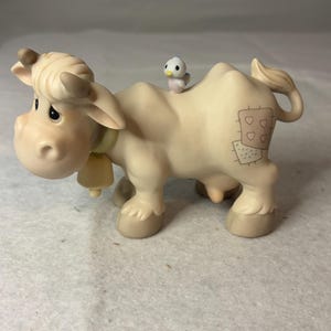 May include: A matte cream-colored cow figurine with brown horns, a yellow bell collar, and a small blue and white bird perched on its back. The cow features a patch design on its rear flank with three small heart outlines and a dotted pattern.