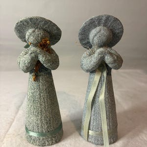 May include: Two gray textured figurines with wide-brimmed hats and long, tapered bodies. One figurine holds a small bundle of dried brown flowers, while the other features a light green ribbon tied around its waist and draped down the front.