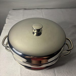 May include: A round, polished stainless steel multi-tiered steamer pot with a matching lid. The lid features a central knob handle, and the pot includes two side handles. The reflective surface shows a subtle distortion of the surrounding environment.