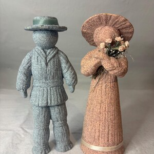 May include: Two textured figurines standing side by side. The left figure is blue, wearing a hat with a green ribbon and a collared jacket. The right figure is peach-colored, wearing a wide-brimmed hat and holding a small bouquet of light pink flowers.