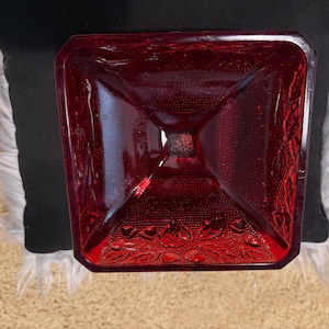 May include: A square-shaped, deep red glass bowl featuring a textured surface with a raised holly leaf and berry pattern along the edges. The glass is translucent, allowing light to pass through, and the design includes a stippled texture.
