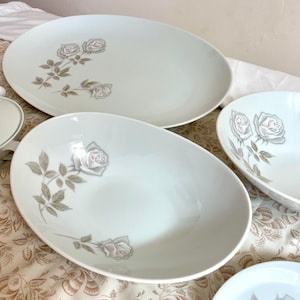 May include: A collection of white porcelain serving dishes with a floral rose design. The set includes oval platters and bowls, each decorated with a gray and pink rose motif. The dishes are arranged on a patterned fabric.