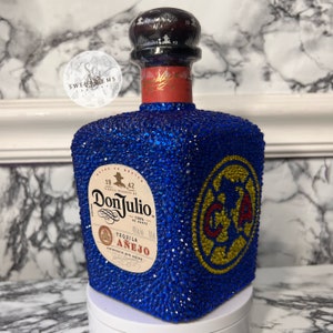 May include: A blue, crystal-covered bottle of Don Julio Añejo tequila with a gold and red logo on the front. The bottle is decorated with a blue and gold soccer team logo.