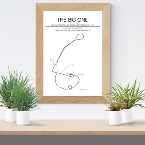 May include: A black and white line drawing of a race track called "The Big One" located in Blackpool, UK. The track is a simple oval shape with a small loop at the top. The drawing is framed in a light wood frame and is hanging on a white wall.