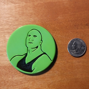 May include: A green 3D printed round coaster with a black silhouette of a person's head and shoulders. The person is bald and has a serious expression.