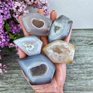 May include: Five agate crystal geodes in various shades of gray, brown, and white. The geodes are all roughly the same size and shape, with a pointed end and a rounded base. They are all polished and have a smooth, shiny surface.