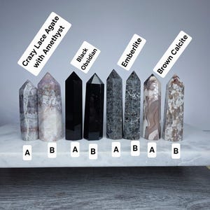 May include: Collection of polished crystal towers, including Crazy Lace Agate with Amethyst, Black Obsidian, Emberlite, and Brown Calcite. Each tower is labeled with its name. The crystals are displayed on a white surface.