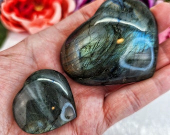 May include: Two heart-shaped labradorite crystals, one larger than the other, with iridescent blue and green hues. The crystals are held in a hand.