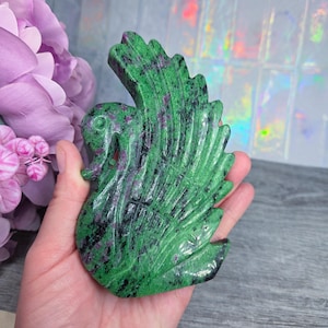 May include: A carved stone swan figurine in shades of green, black, and red. The swan is held in a hand, showcasing the intricate details of the carved feathers and body. The stone has a polished finish.
