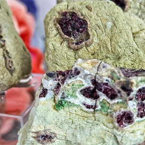 May include: Two pieces of rough, green and white stone with red and purple crystals embedded in the surface.