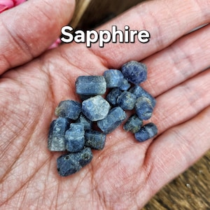 May include: A collection of small, rough, blue sapphire gemstones. The gemstones are in various shapes and sizes, and they are all clustered together in a hand.