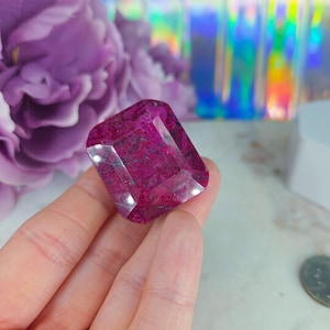 May include: A large, rectangular, faceted gemstone with a deep magenta color. The gem is held in a hand, with a blurred purple flower and iridescent background. The gem's facets catch the light, creating a sparkling effect.
