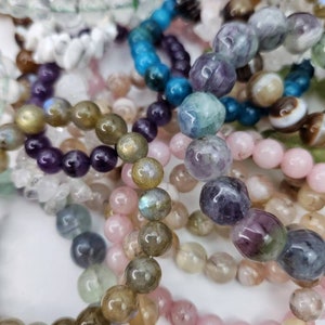 May include: A collection of beaded bracelets in various colors and textures. The bracelets feature a variety of gemstones, including pink, purple, blue, and green beads. Some beads have a polished, glossy finish, while others appear more matte.