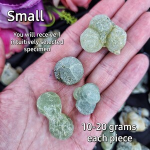 May include: A hand holding a variety of green, translucent, smooth, round, and irregularly shaped stones. The stones are described as "Small" and weigh between 10-20 grams each. The text "You will receive 1 intuitively selected specimen" is also visible.