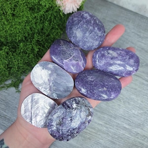 May include: A hand holds several polished, oval-shaped stones in shades of purple and gray. The stones vary in size and have a smooth, reflective surface. The background includes green moss and a few pink flowers, suggesting a natural setting.