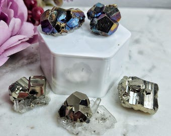 Puede incluir: Un surtido de cristales iridiscentes y metálicos. Dos cristales con tonos azules, morados y dorados se encuentran sobre una caja hexagonal blanca. Varios cristales de pirita se exhiben sobre una superficie blanca.