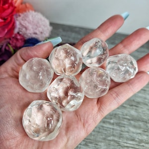 May include: A hand holds several clear quartz tumbled stones. The stones are smooth, rounded, and translucent, with a slightly cloudy appearance. The quartz crystals vary in size and shape, reflecting light.