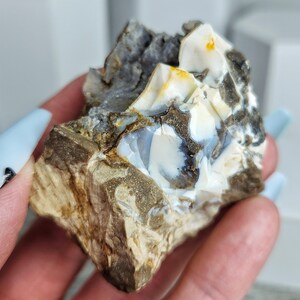 May include: A rough, natural stone with a mix of colors. The stone features areas of white, gray, brown, and hints of yellow. The texture appears to be layered and crystalline, with a raw, unpolished appearance.