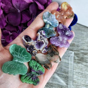 May include: A collection of carved stone butterfly figurines in various colors, including green, purple, and brown. The butterflies are held in a hand, with purple flowers in the background. The figurines are detailed and appear to be made of different types of stone.