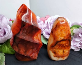 May include: Two Carnelian crystal freeform carvings. The larger carving is a tall, pointed shape with a red, orange, and white banded pattern. The smaller carving is a rounded, smooth shape with a similar banded pattern.