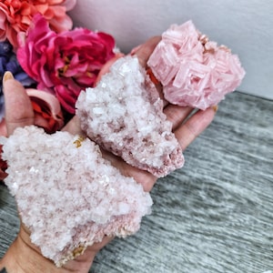 May include: Three pink Himalayan salt crystals of varying sizes and shapes. The crystals are clustered together and have a rough, textured surface.