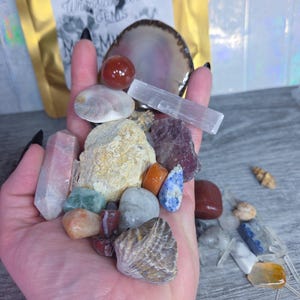May include: A collection of various gemstones and crystals, including a rose quartz crystal, a selenite wand, and several polished stones in shades of red, blue, orange, and gray. A seashell and a small bag with the text "Whimsical Gems" are also visible.