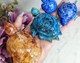 May include: A collection of carved gemstone turtle figurines in shades of blue and amber. The turtles vary in size and detail, showcasing the natural patterns and colors of the stones. The figurines are displayed on a hand.