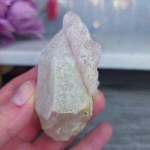 May include: A light pink quartz crystal with a rough, textured surface. The crystal has a pointed tip and a layered appearance, with a lighter pink hue at the top. The base of the crystal is a pale, off-white color. The crystal is a natural, raw gemstone.