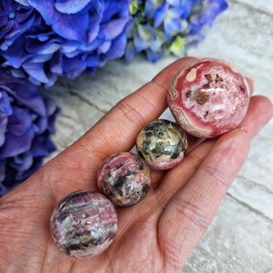 May include: Four pink and grey gemstone spheres of varying sizes, held in a hand. The spheres are smooth and polished, with a variety of patterns and textures.