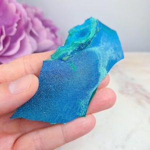 May include: A vibrant blue and green mineral specimen with a rough, irregular shape. The stone displays a mix of deep blue and lighter turquoise hues, with areas of textured, crystalline formations. The mineral is held in a hand, with a blurred purple flower in the background.