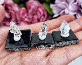 May include: Three clear quartz crystals on black square bases. The crystals are pointed and have a natural, rough texture. The bases are made of polished black stone.
