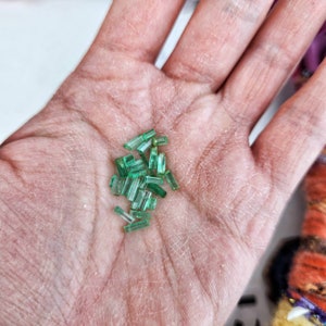 May include: A collection of small, green emerald gemstones in a variety of shapes and sizes. The gemstones are arranged in a pile on a person's hand.