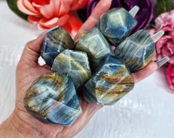 May include: A collection of seven blue and gold banded, faceted, polished, gemstone tumbled stones. The stones are in the palm of a hand.