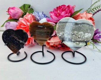 May include: Three heart-shaped polished stones on black metal stands. The stones are brown, black and white with natural patterns. The stones are displayed against a background of pink and purple flowers.
