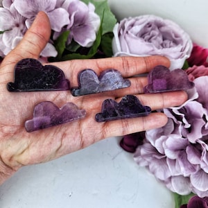 May include: Five purple amethyst crystal cloud shaped stones on a hand. The stones are various shades of purple and have a cloudy, translucent appearance.