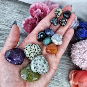 May include: A collection of heart-shaped polished stones in various colors, including amethyst, green aventurine, carnelian, and jasper. The stones are arranged on a hand.