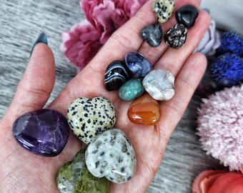 May include: A collection of heart-shaped polished stones in various colors, including amethyst, green aventurine, carnelian, and jasper. The stones are arranged on a hand.