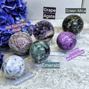 May include: A collection of polished crystal spheres, including green mica, grape agate, pink tourmaline, emerald, hackmanite, and stichtite, also known as atlantisite. The spheres are displayed on small stands.