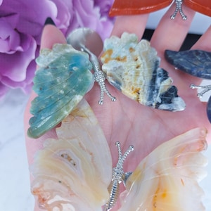 May include: A collection of gemstone butterfly figurines with silver-colored metal accents. The butterflies feature wings in various colors, including blue, beige, and orange. The wings have intricate patterns and textures, showcasing the natural beauty of the stones.