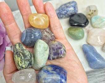 May include: A collection of polished gemstones in various colors, including yellow, blue, green, purple, and clear. The stones are held in a hand, showcasing their different shapes and textures. The gemstones are likely used for decorative or spiritual purposes.