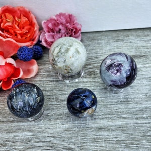 May include: Four polished crystal spheres on display stands. The spheres are various colors, including white, blue, and purple. The spheres are on a gray wooden surface.