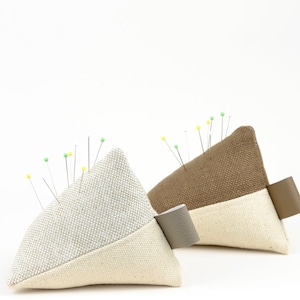 May include: Two triangular pincushions with different coloured fabric tops. The pincushions are filled with stuffing and have a white fabric base. The pincushions are filled with pins with coloured heads.