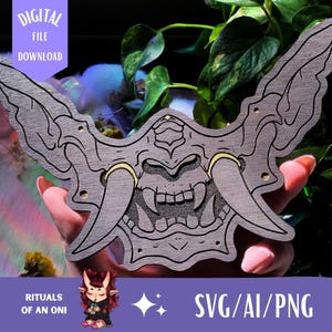 May include: A gray wooden Oni mask with black outlines and gold accents. The mask has large horns and a fierce expression. Text on the image reads "DIGITAL FILE DOWNLOAD" and "SVG/AI/PNG". The image is of a digital file for a craft project.