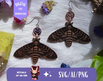 May include: A pair of wooden earrings featuring a black and brown moth design with a skull detail. The earrings are hanging from silver hooks with a pink and silver crescent moon charm.