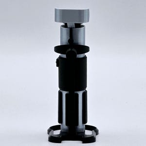 May include: A silver and black lightsaber prop with a black handle and a silver blade. The lightsaber is on a black stand.