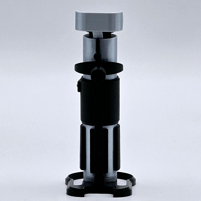 May include: A silver and black lightsaber prop with a black handle and a silver blade. The lightsaber is on a black stand.