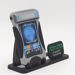 Może przedstawiać: Replika Star Trek Nemesis Starfleet Tricorder. Urządzenie jest szare i czarne, z niebieskim ekranem wyświetlającym siatkę celowniczą. Podstawa jest czarna, a na wyświetlaczu widnieje napis "MONOMOLECULAR SCAN RATE". Osobna czarna tabliczka głosi "STAR TREK NEMESIS STARFLEET TRICORDER".