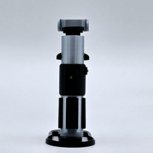 May include: A silver and black lightsaber prop with a green light on the handle. The lightsaber is on a black stand.