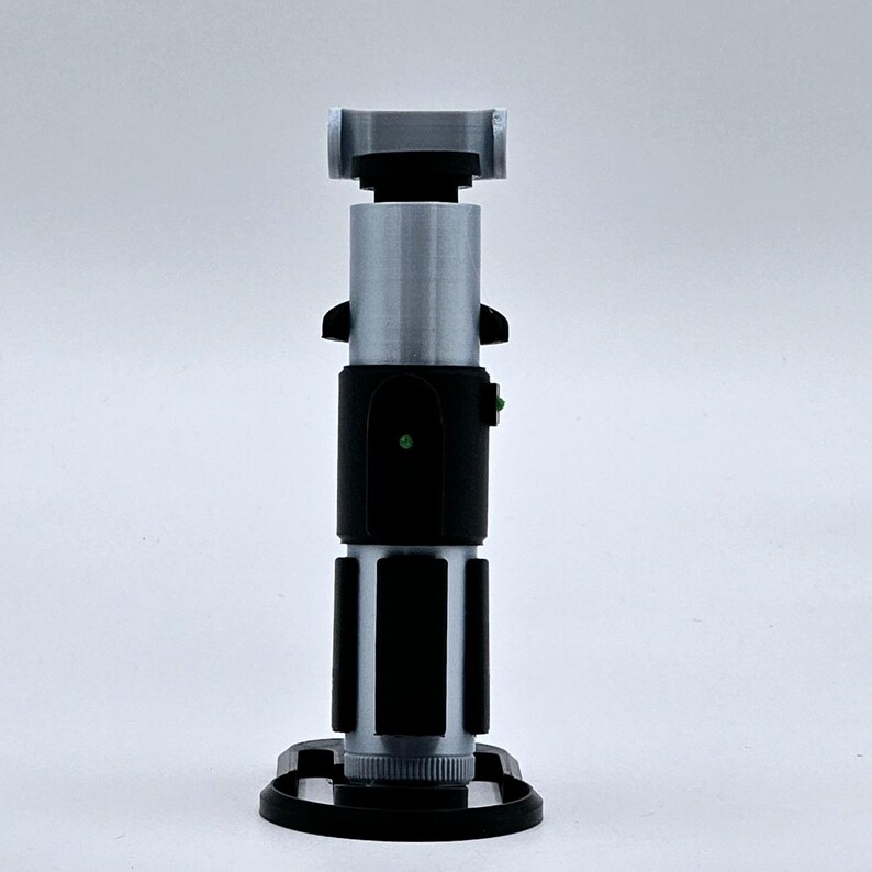 May include: A silver and black lightsaber prop with a green light on the handle. The lightsaber is on a black stand.